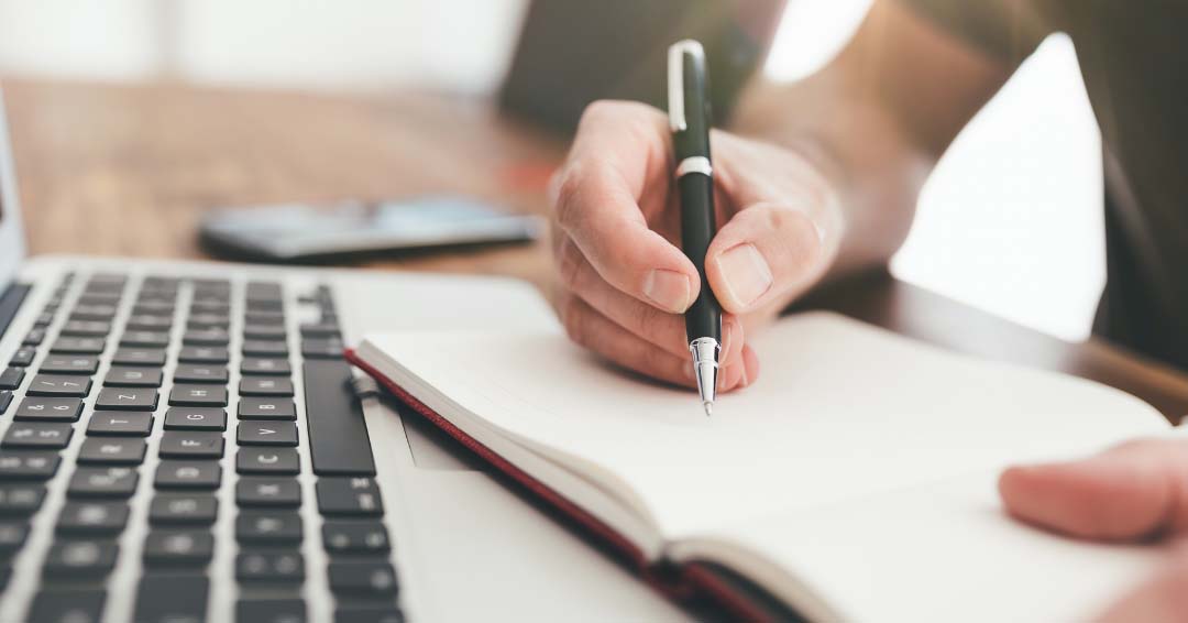 person taking notes on a pad next to a computer keyboard